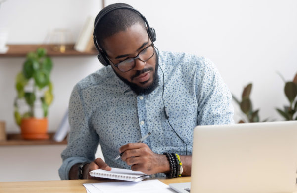 Focused african business man in headphones writing notes in notebook watching webinar video course, serious black male student looking at laptop listening lecture study online on computer e learning